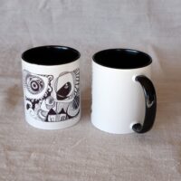 Sur cette photo il y 2 Mugs en céramique blanche à anse et intérieur noirs. Sur l'un est imprimé un dessin d'artiste en noir qui représente dans une forme rectangulaire des maisons, des fleurs, des vagues, des spirales très stylisées contenant beaucoup de détails. L'autre mug est positionné différemment du côté de son anse noire. Nous voyons que le reste de la tasse est blanc et apparaît sur le côté gauche du motif une partie du dessein décrit ci-dessus.